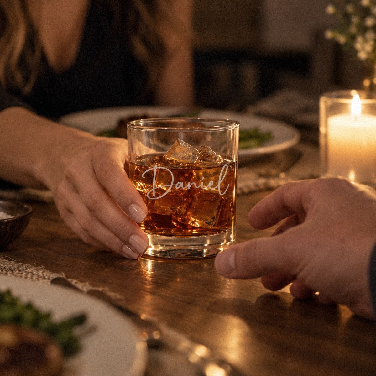 A woman slides a personalized whiskey glass engraved Daniel across a candlelit dinner table to him, one of the romantic valentine gifts for him