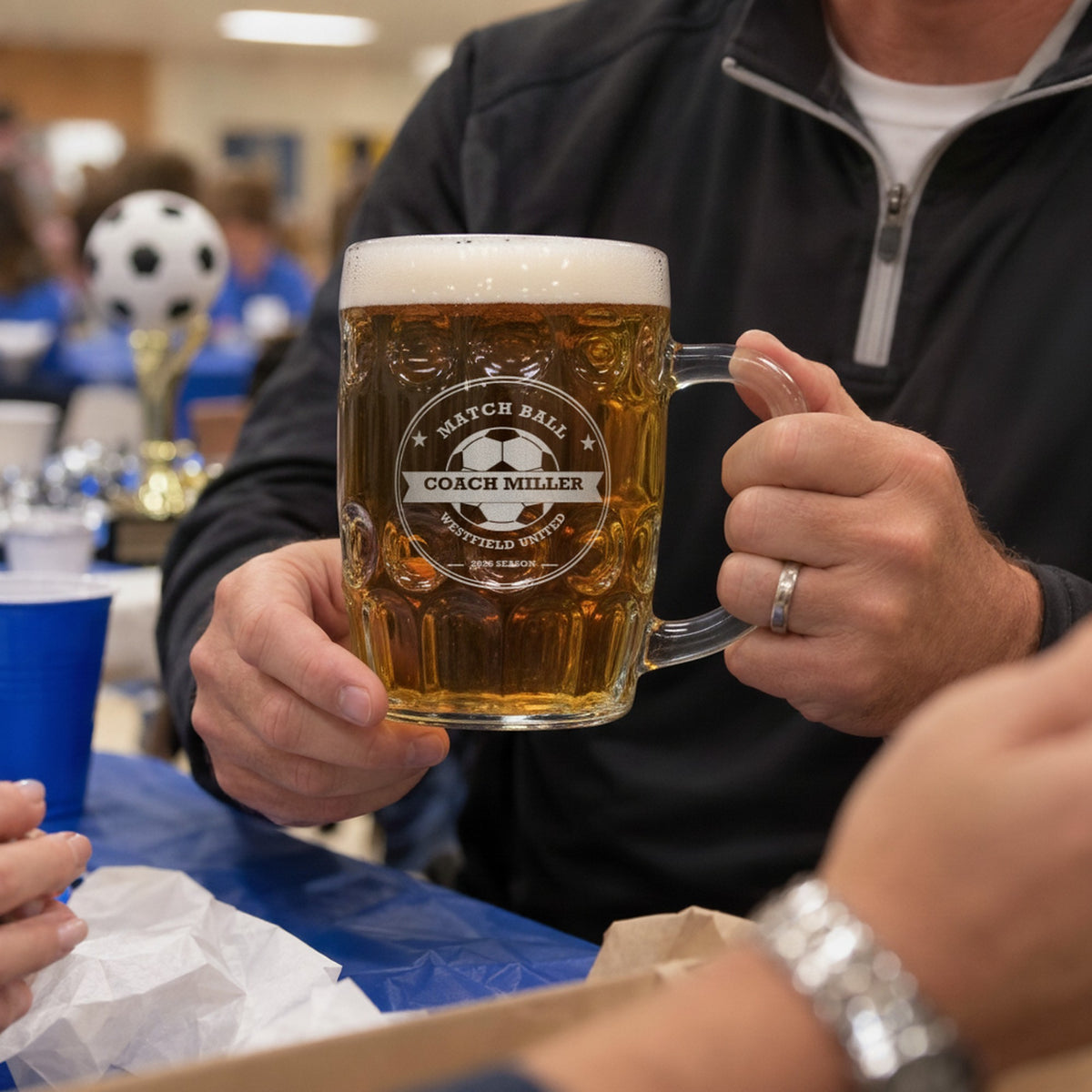 Soccer coach holding the personalized Match Ball beer stein at an end-of-season team banquet, with a soccer trophy in the background