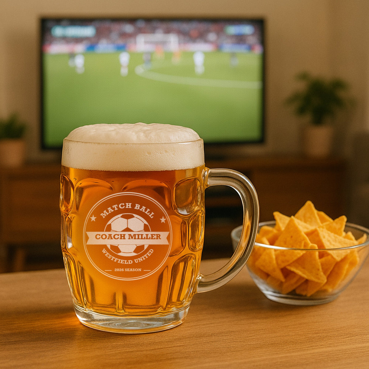 Custom soccer coach beer stein of lager on a living room table beside a bowl of chips, with a soccer match playing on the TV behind it