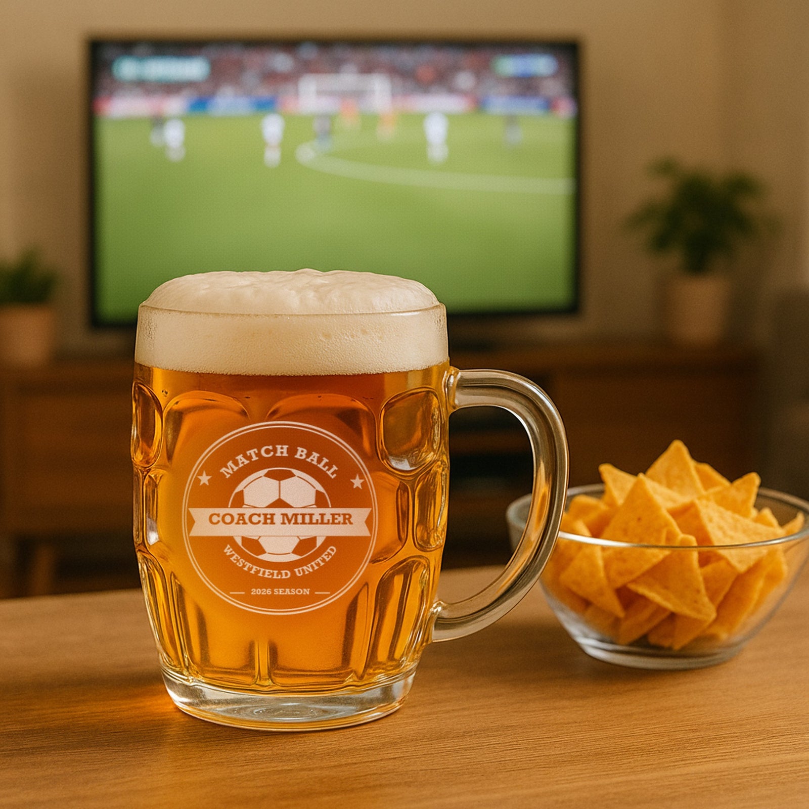 Custom soccer coach beer stein of lager on a living room table beside a bowl of chips, with a soccer match playing on the TV behind it