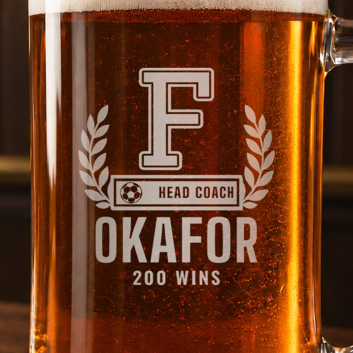 Close-up of the frosted engraving on the soccer coach letterman beer stein, a varsity letter with laurel and a soccer ball COACH bar: F, Head Coach, Okafor, 200 Wins