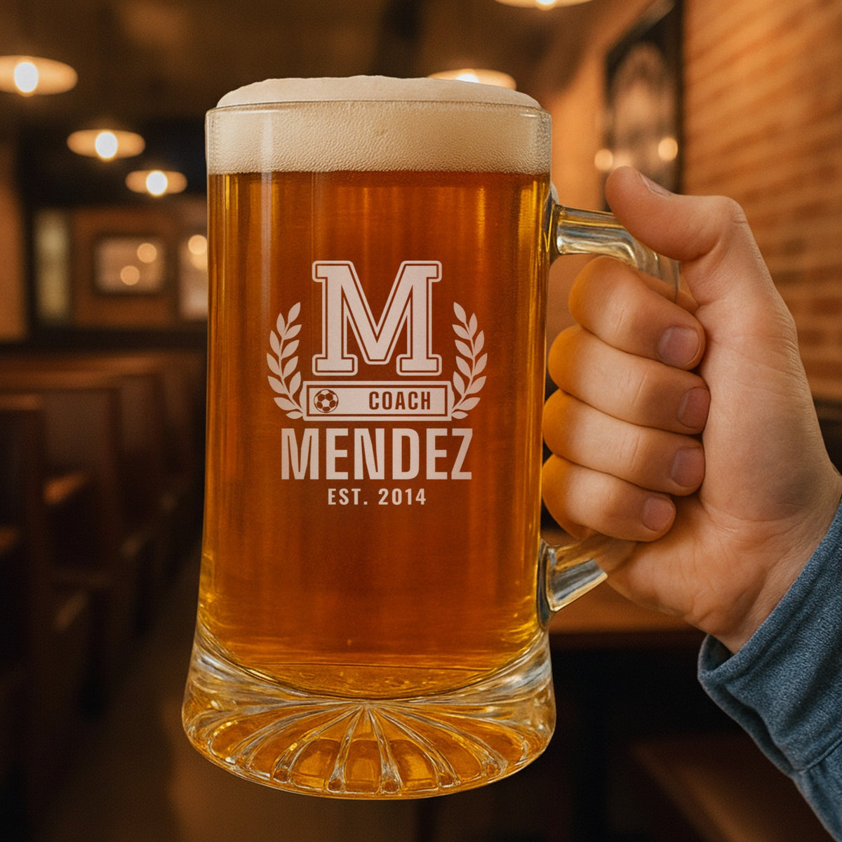 Hand holding the 25 oz personalized soccer coach letterman beer stein in a pub, engraved with a varsity letter with laurel and a soccer ball COACH bar: M, Coach, Mendez, Est. 2014