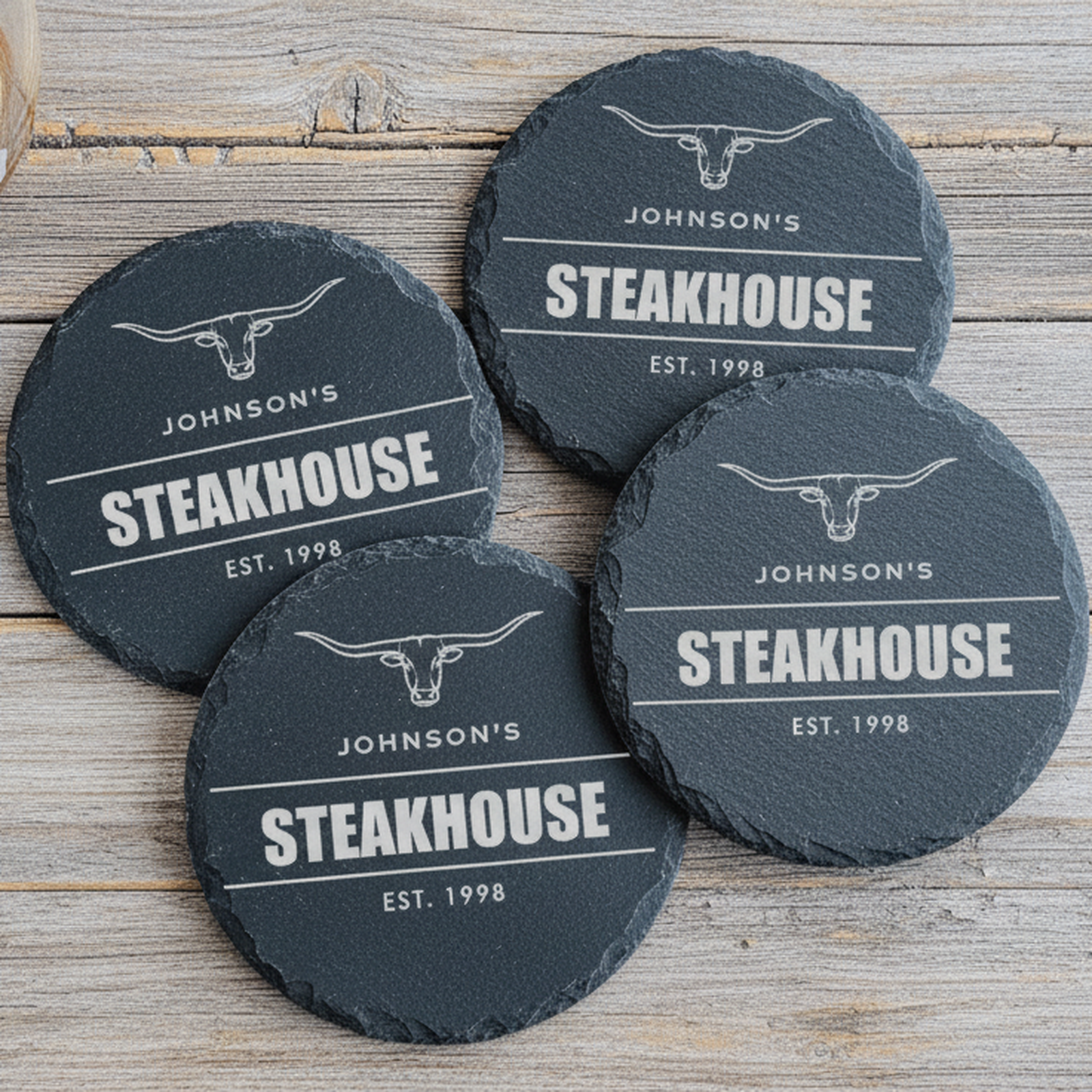 Set of four personalized steakhouse slate coasters engraved with a longhorn emblem, family name, big word, and established year, styled on rustic wood with a whiskey glass