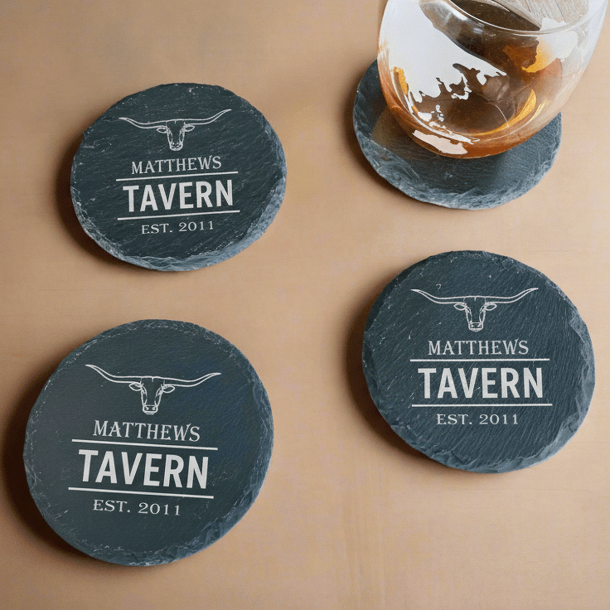 Personalized steakhouse slate coasters engraved with a longhorn emblem, family name, big word, and established year, with a whiskey glass resting on a matching coaster