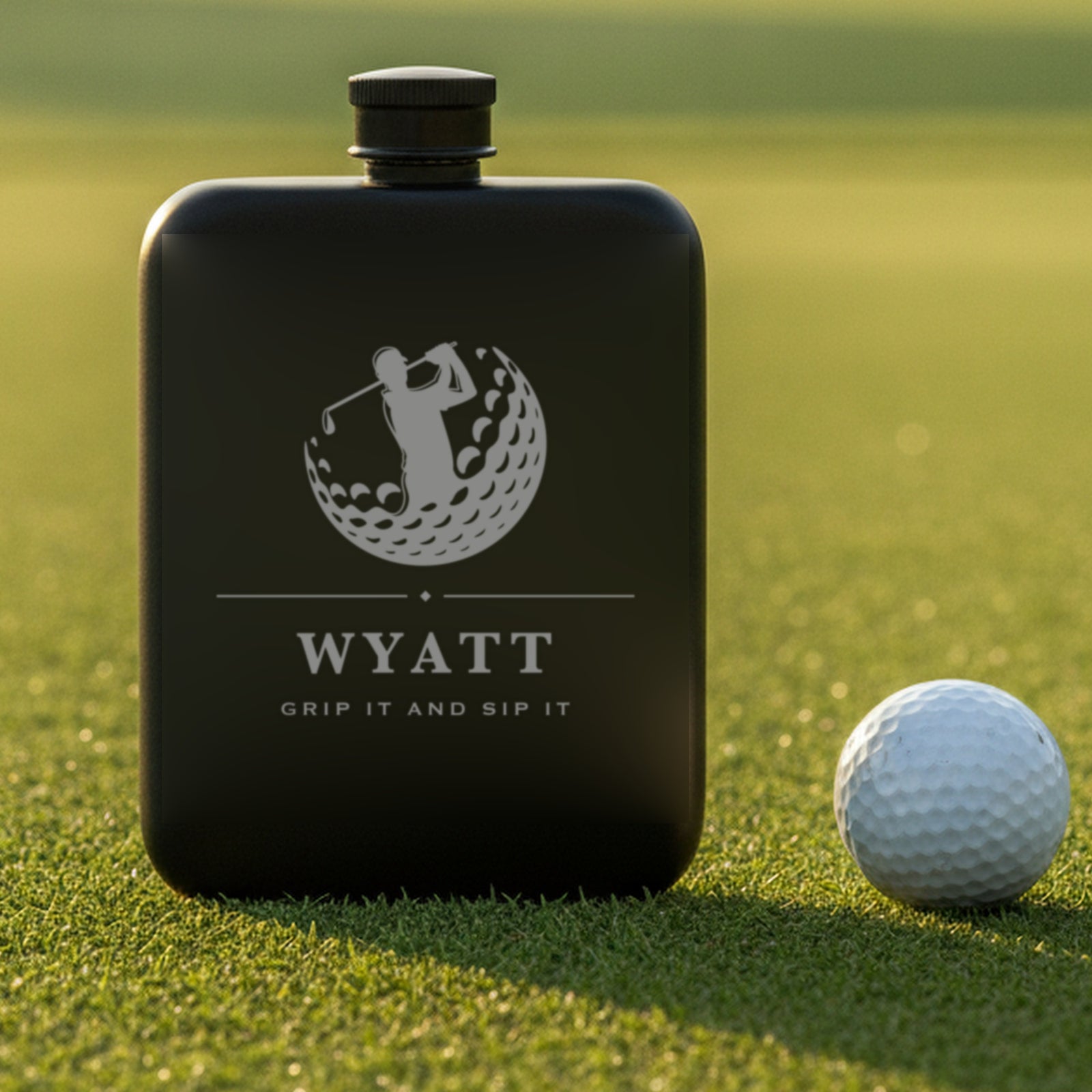 Engraved golf flask reading Grip It And Sip It standing on putting green turf beside a white golf ball in morning light.