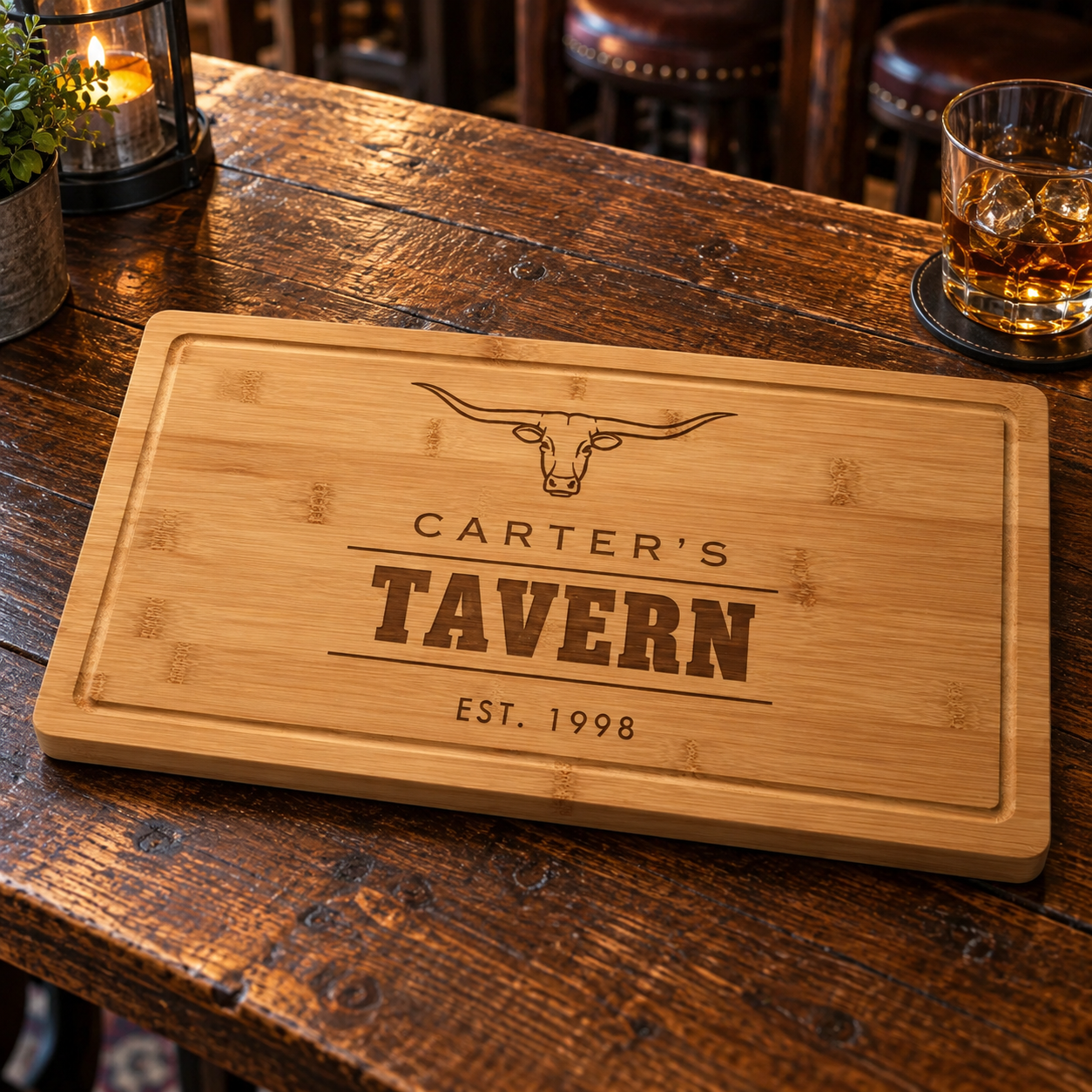 Bamboo cutting board with engraved longhorn design and sample text reading "Carter's Tavern Est. 1998" on dark wood table with whiskey glass