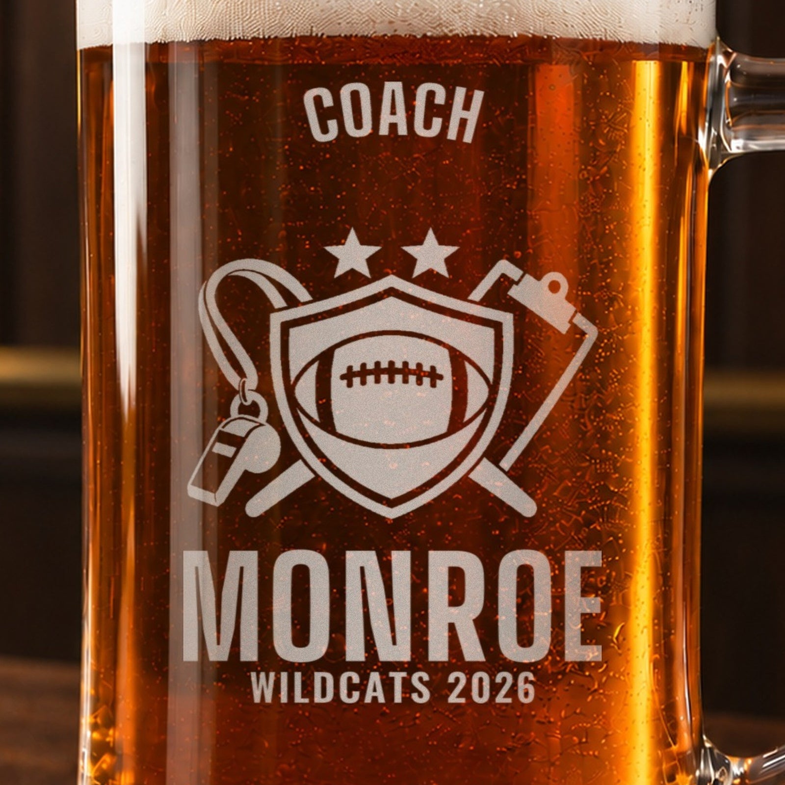 Close-up of the frosted engraving on the thanks coach football beer stein, a shield with a football, a whistle and a clipboard: Coach, Monroe, Wildcats 2026