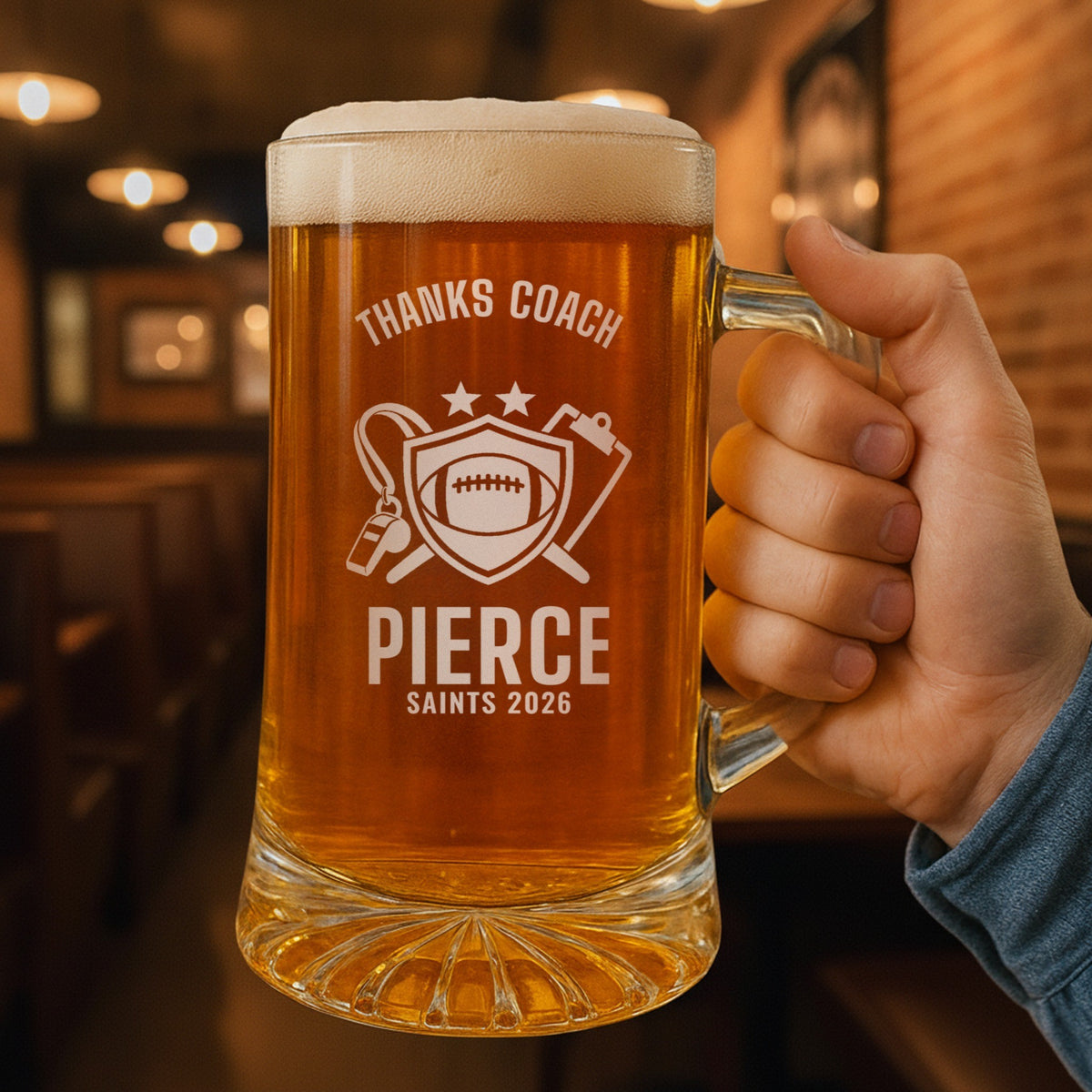 Hand holding the 25 oz personalized thanks coach football beer stein in a pub, engraved with a shield with a football, a whistle and a clipboard: Thanks Coach, Pierce, Saints 2026