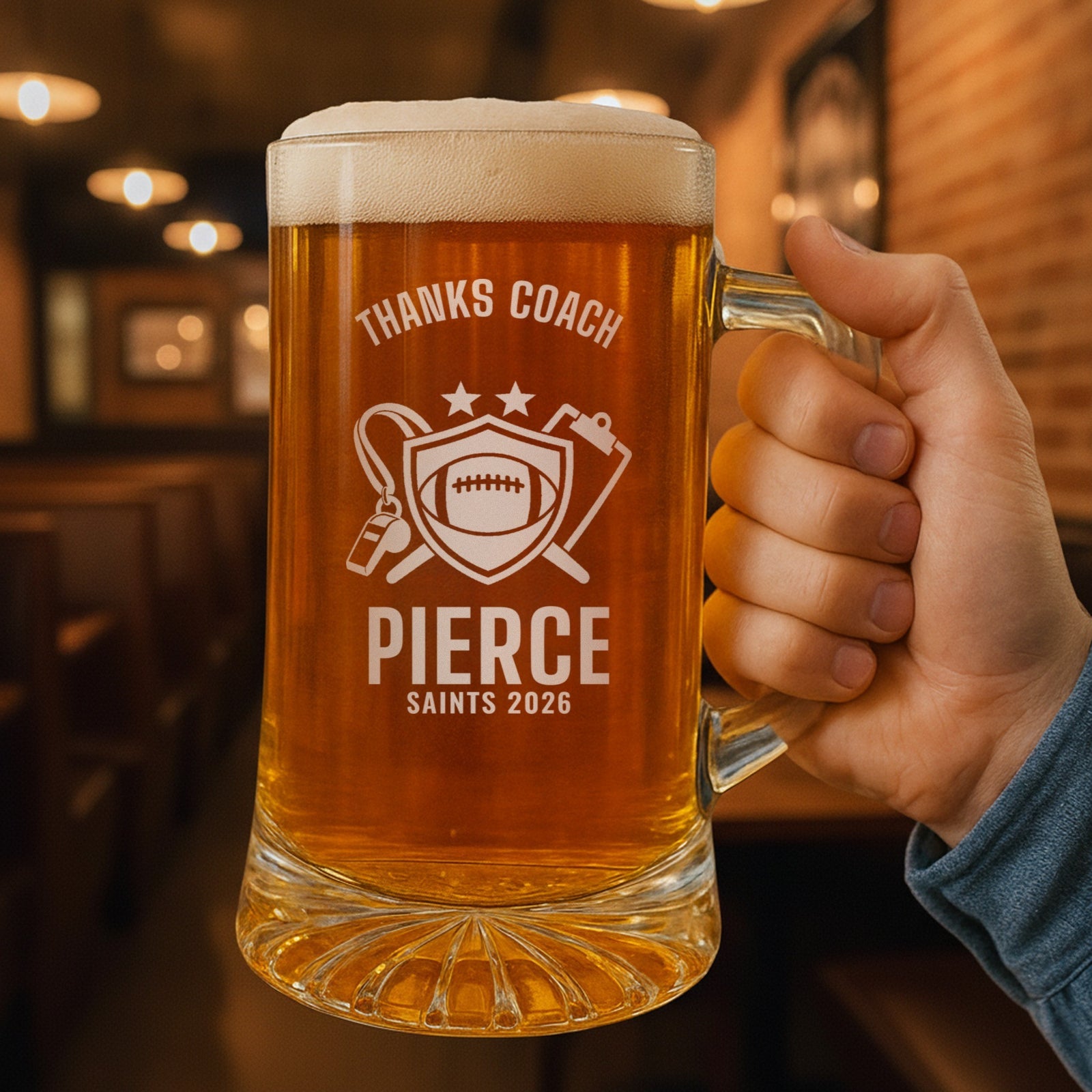 Hand holding the 25 oz personalized thanks coach football beer stein in a pub, engraved with a shield with a football, a whistle and a clipboard: Thanks Coach, Pierce, Saints 2026
