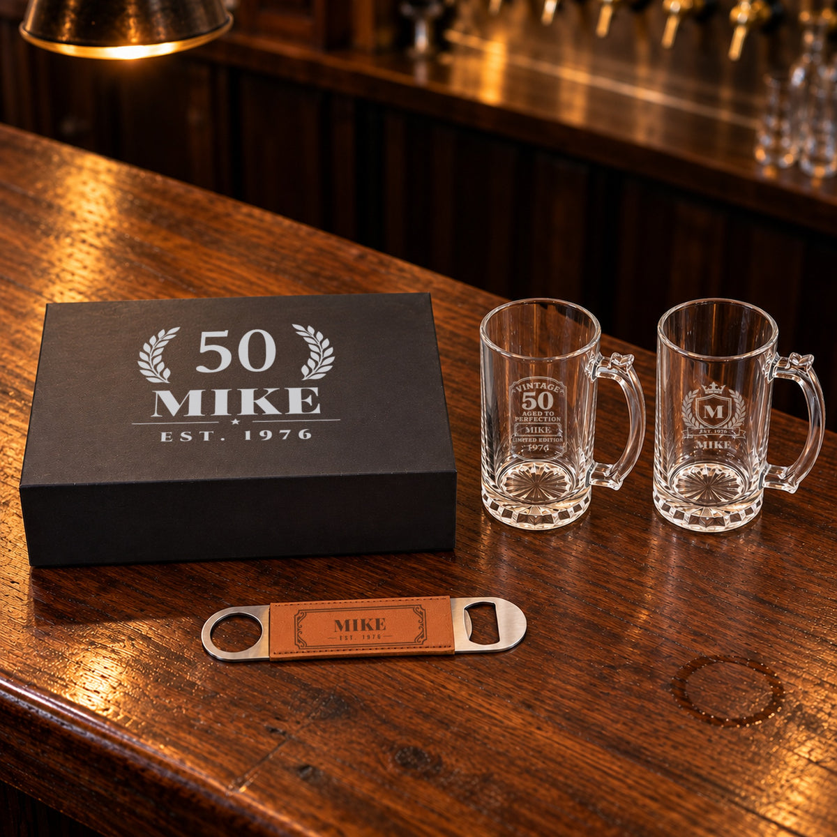 50th birthday beer gift set on a bar: box lid engraved 50 in a laurel with Mike and EST. 1976, a Vintage 50 mug, a crest mug and an opener
