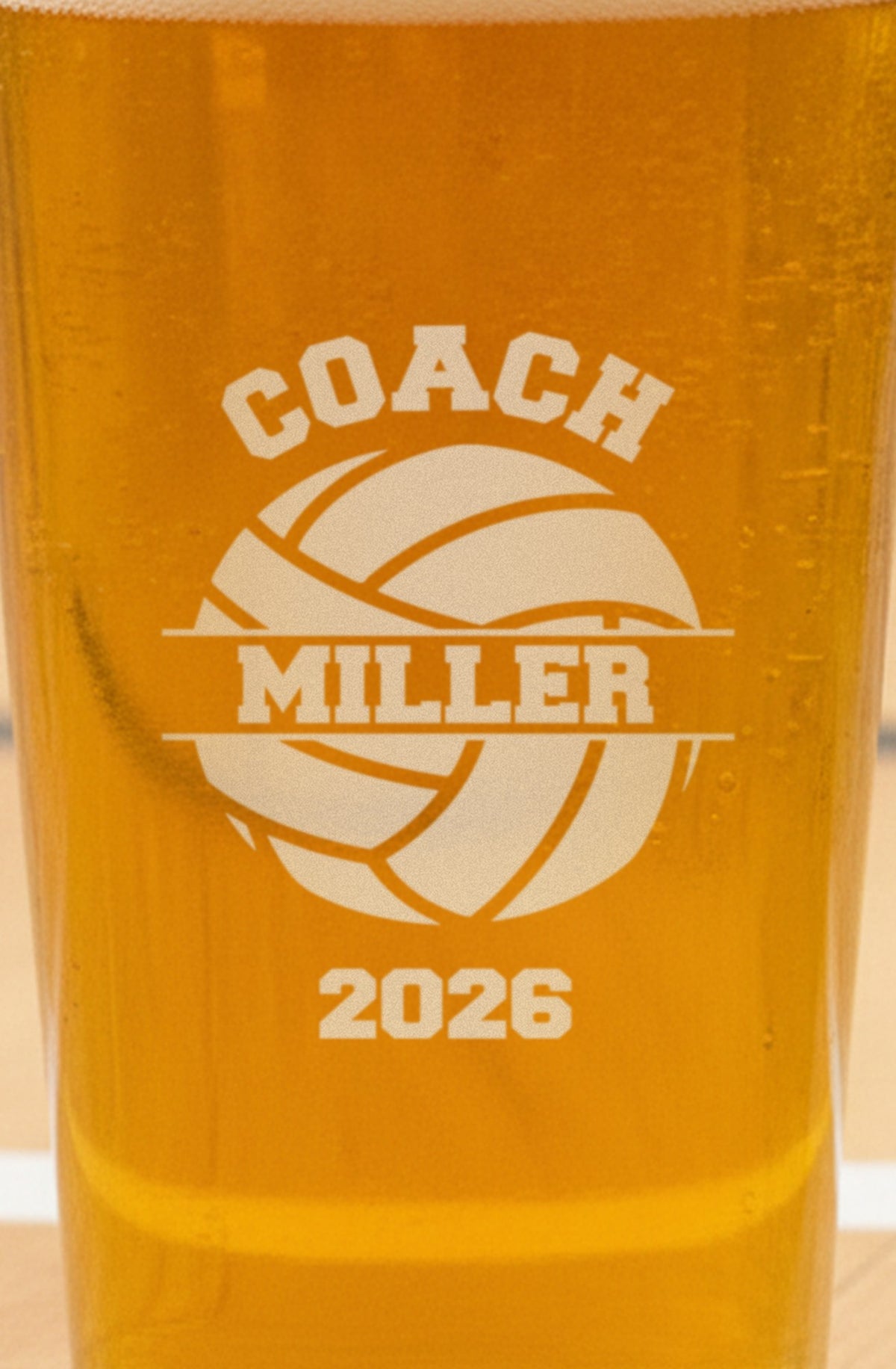 Close-up of the frosted engraving on the volleyball coach pint glass: COACH arched over a volleyball, Miller in the banner and the year 2026