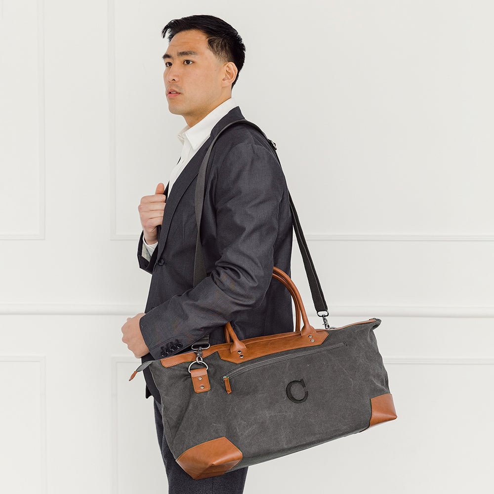 Man in a dark suit carrying a charcoal canvas weekender on its shoulder strap, the initial C embroidered in black on the front of the bag of the personalized travel bag