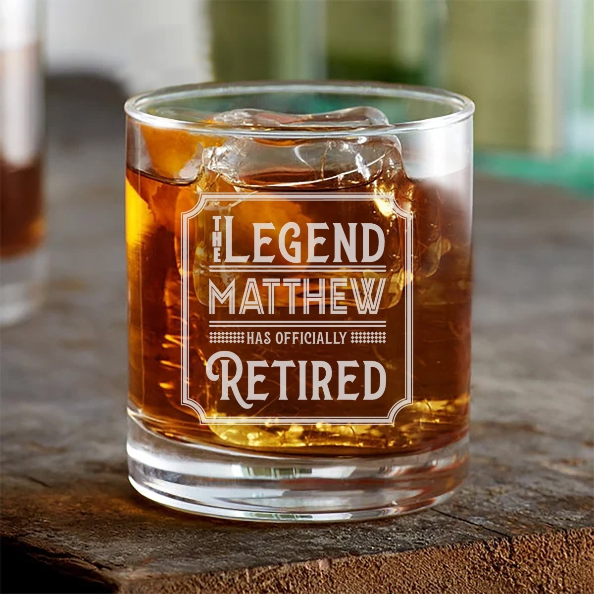 Original photo of the retirement whiskey glass engraved THE LEGEND MATTHEW HAS OFFICIALLY RETIRED, a personalized rocks glass over ice on a wood table.