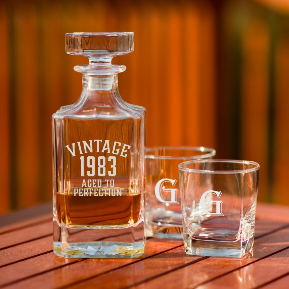 Vintage Birthday Decanter Set on a wood deck railing: glass decanter engraved VINTAGE 1983 AGED TO PERFECTION beside two rocks glasses engraved G