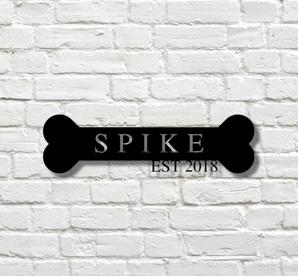 Black laser cut steel dog bone sign with the name SPIKE and EST 2018 hanging alone on a white painted brick wall, a custom dog name sign