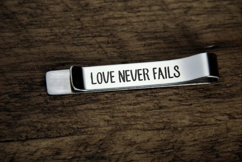 Silver custom tie clip engraved LOVE NEVER FAILS in block capitals on the front, lying on a dark wood table