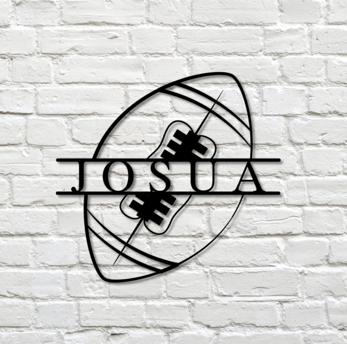 Black metal football sign with JOSUA cut in solid capitals across the middle of the ball, hanging alone on a white painted brick wall, a custom football wall sign