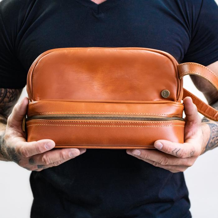 Man holding a tan leather toiletry bag in both hands in front of a black T-shirt, the leather wrist strap on the right, a man leather toiletry bag