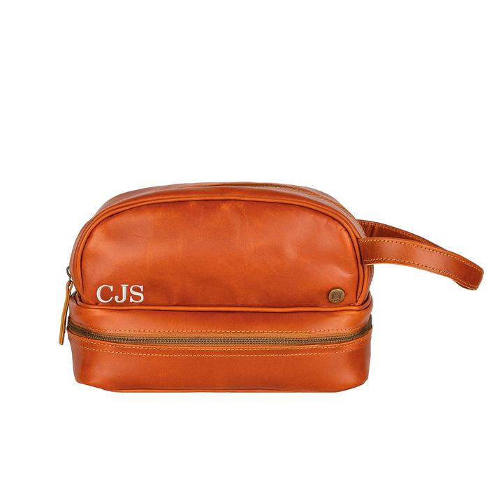Tan personalized leather wash bag with the initials CJS embroidered in cream thread on the front, the wrist strap on the right, on a white background