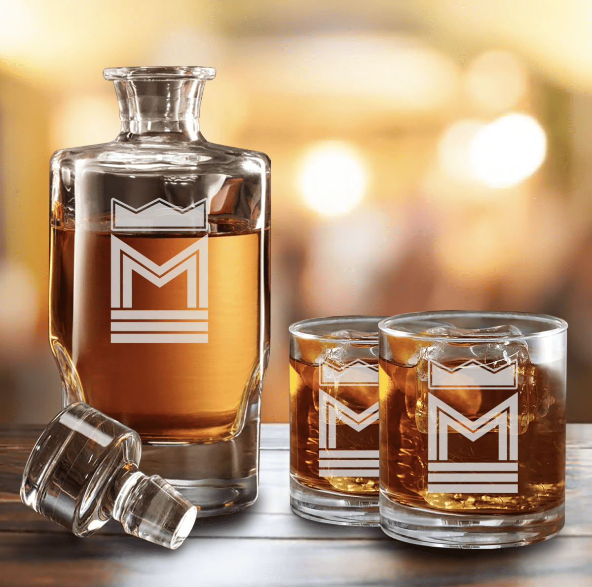 Square glass decanter beside two rocks glasses of bourbon on ice, all engraved with a crown over the letter M, an initial decanter set and unique whiskey gift for men