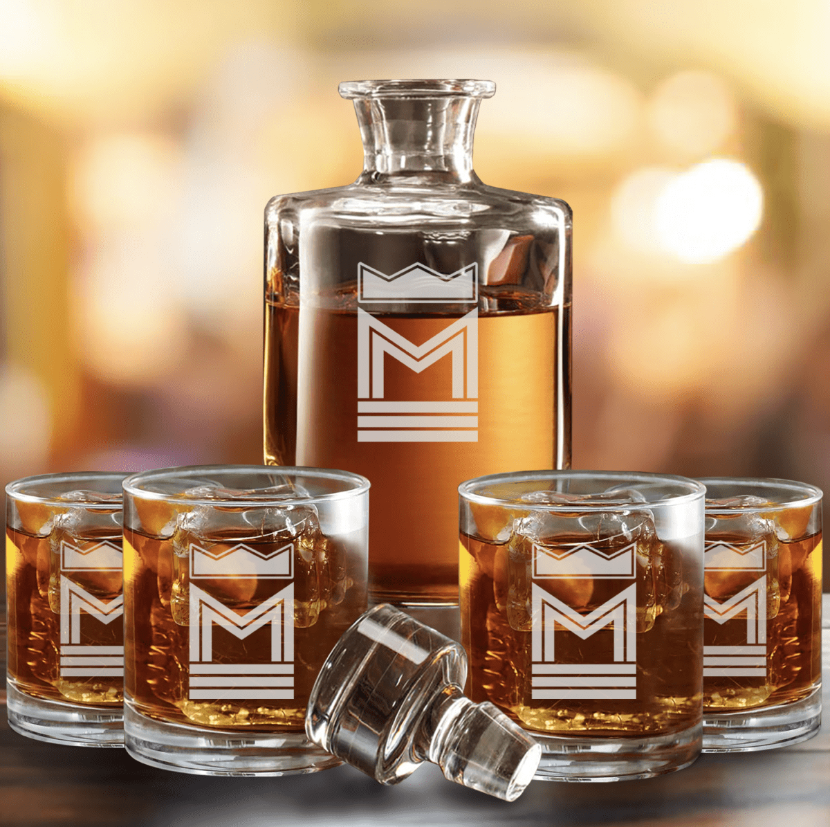 Glass decanter and four rocks glasses of whiskey, each engraved with a crown above a capital M, a personalized initial decanter set with its stopper resting in front